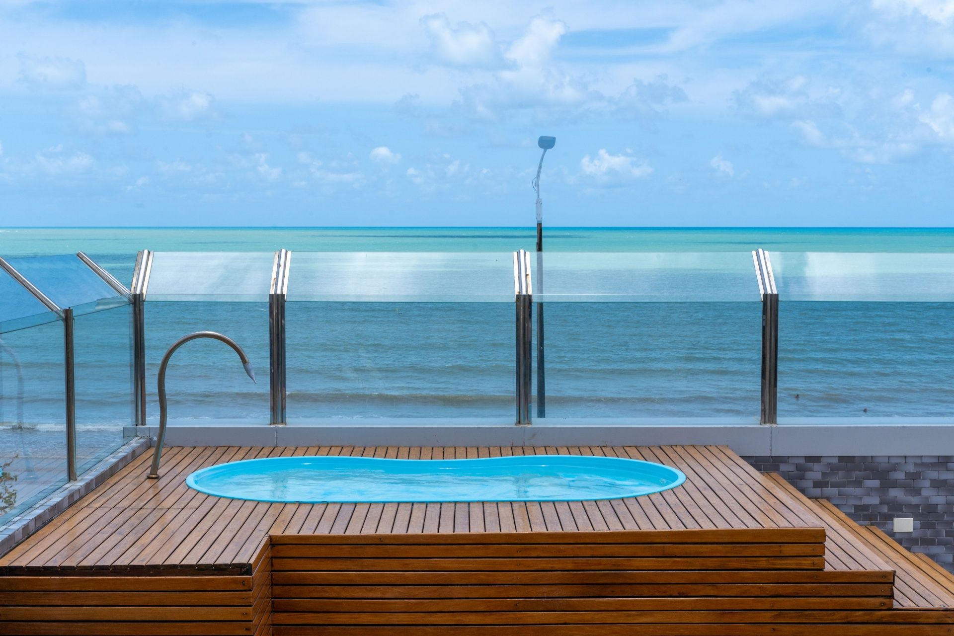 Deck de madeira à beira-mar com uma pequena piscina azul e guarda-corpo de vidro com vista para o oceano.