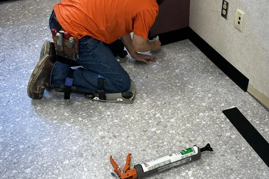 Person kneeling, installing black baseboard trim on a floor. Orange shirt, tool belt, and knee pads are visible.
