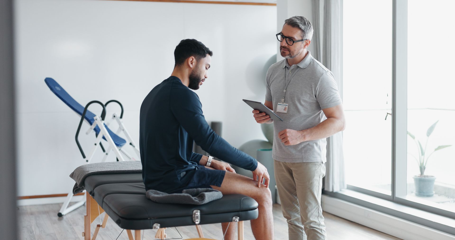 Physical therapist consulting a seated patient in a bright clinic room, with a folded wheelchair nearby