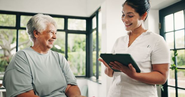 Caregiver smiling with tablet beside seated patient in bright clinic room Caregiver smiling with tablet beside seated patient in bright clinic room