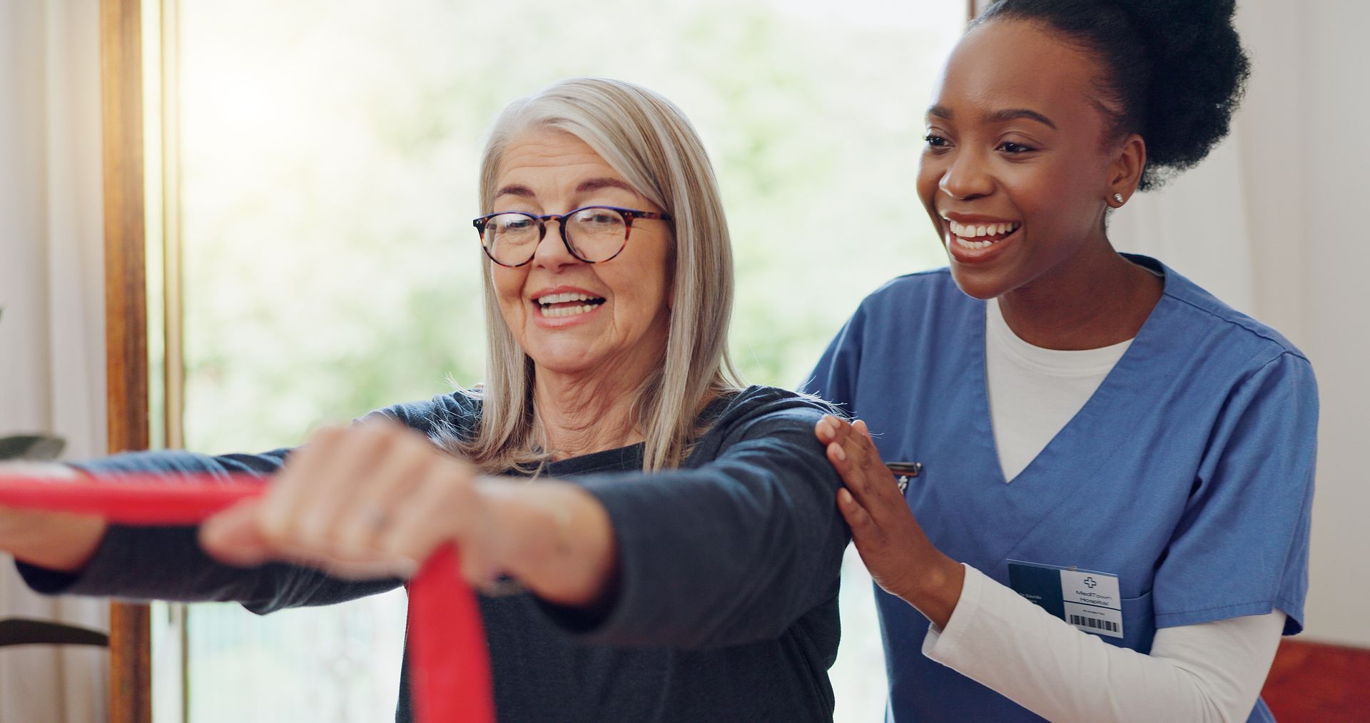 Senior woman doing arm exercise with a smiling caregiver in blue scrubs assisting her indoors Senior woman doing arm exercise with a smiling caregiver in blue scrubs assisting her indoors