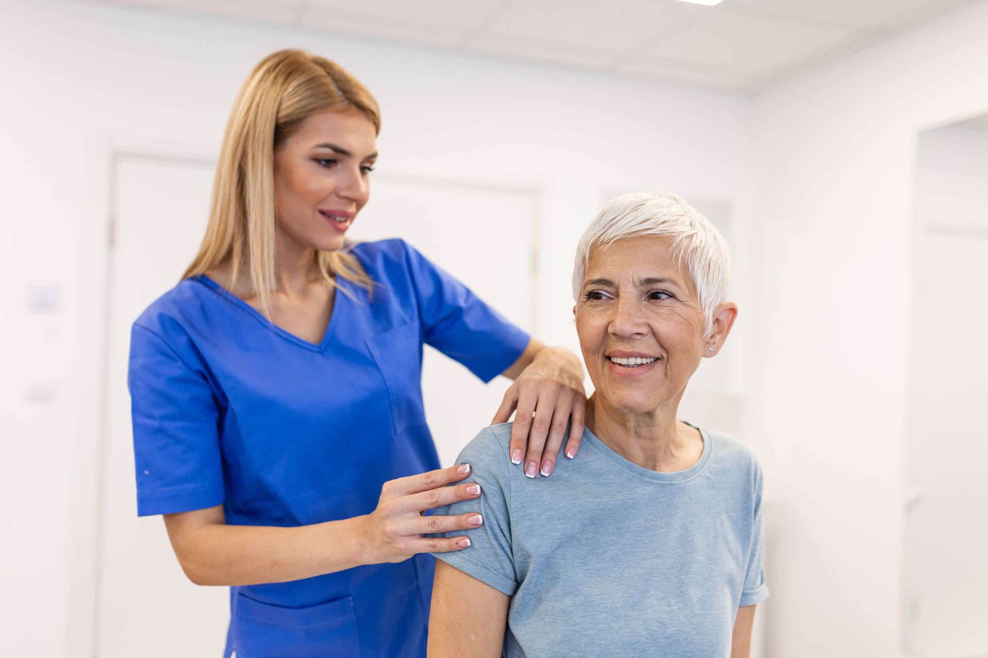 Nurse in blue scrubs smiling and supporting an elderly patient in a bright clinic room