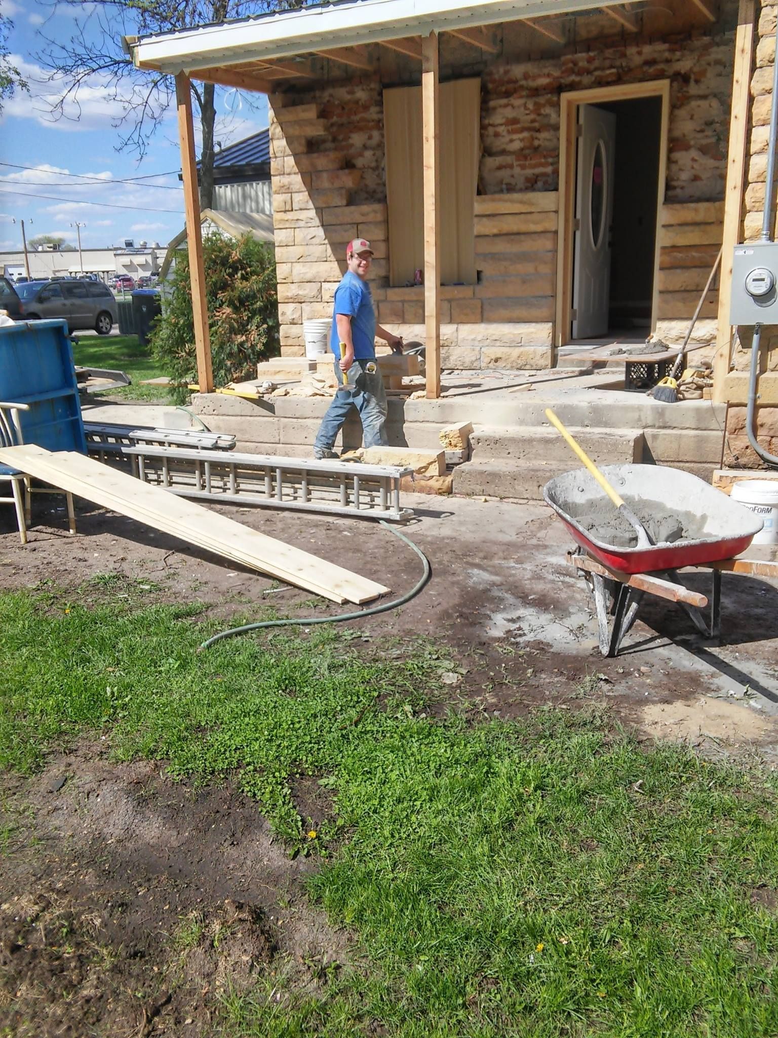 A man is standing in front of a log cabin with a wheelbarrow full of cement.