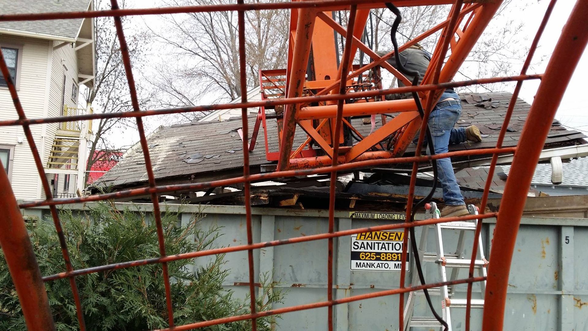 A man on a ladder is working on a crane behind a fence.