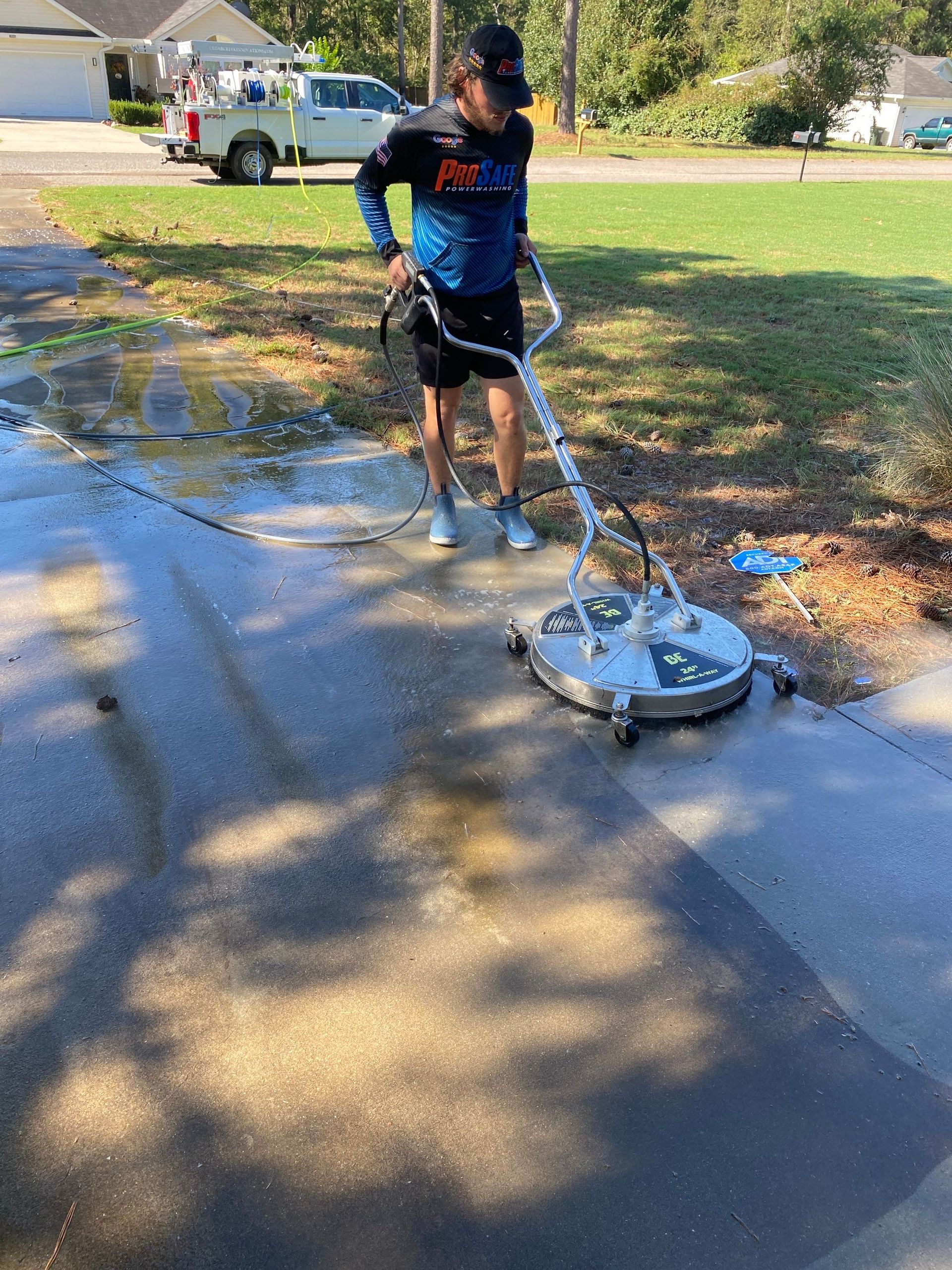 A person in a blue shirt uses a circular pressure washer surface cleaner to clean a concrete driveway on a sunny day.