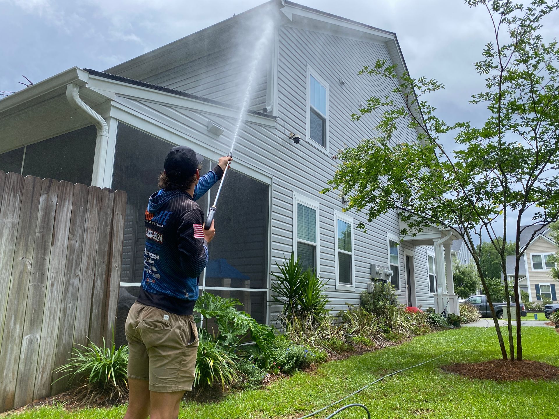 Person power washing siding of a light gray two-story house, spraying water. The setting is a yard with green grass.