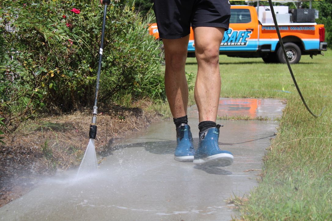 Person power washing a concrete sidewalk outside with a pressure washer, with a truck in the background.