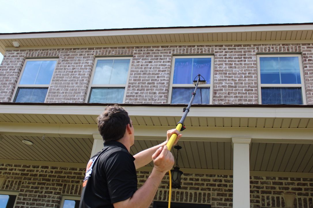 Man washing windows on a two-story house with a long-handled tool under a blue sky.