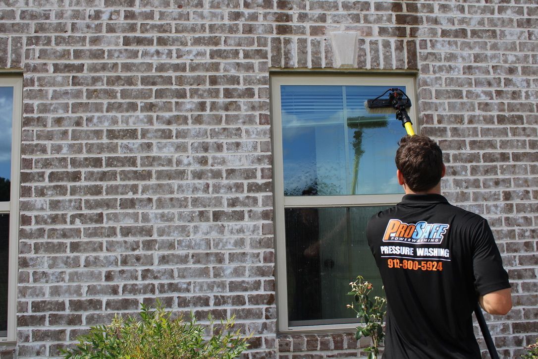 Man cleaning windows on a two-story brick house with a long-handled tool on a sunny day.