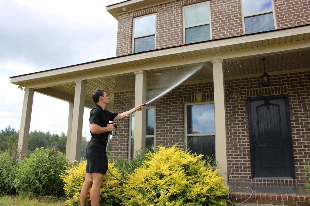 Person power washing a gray house with white trim on a sunny day.