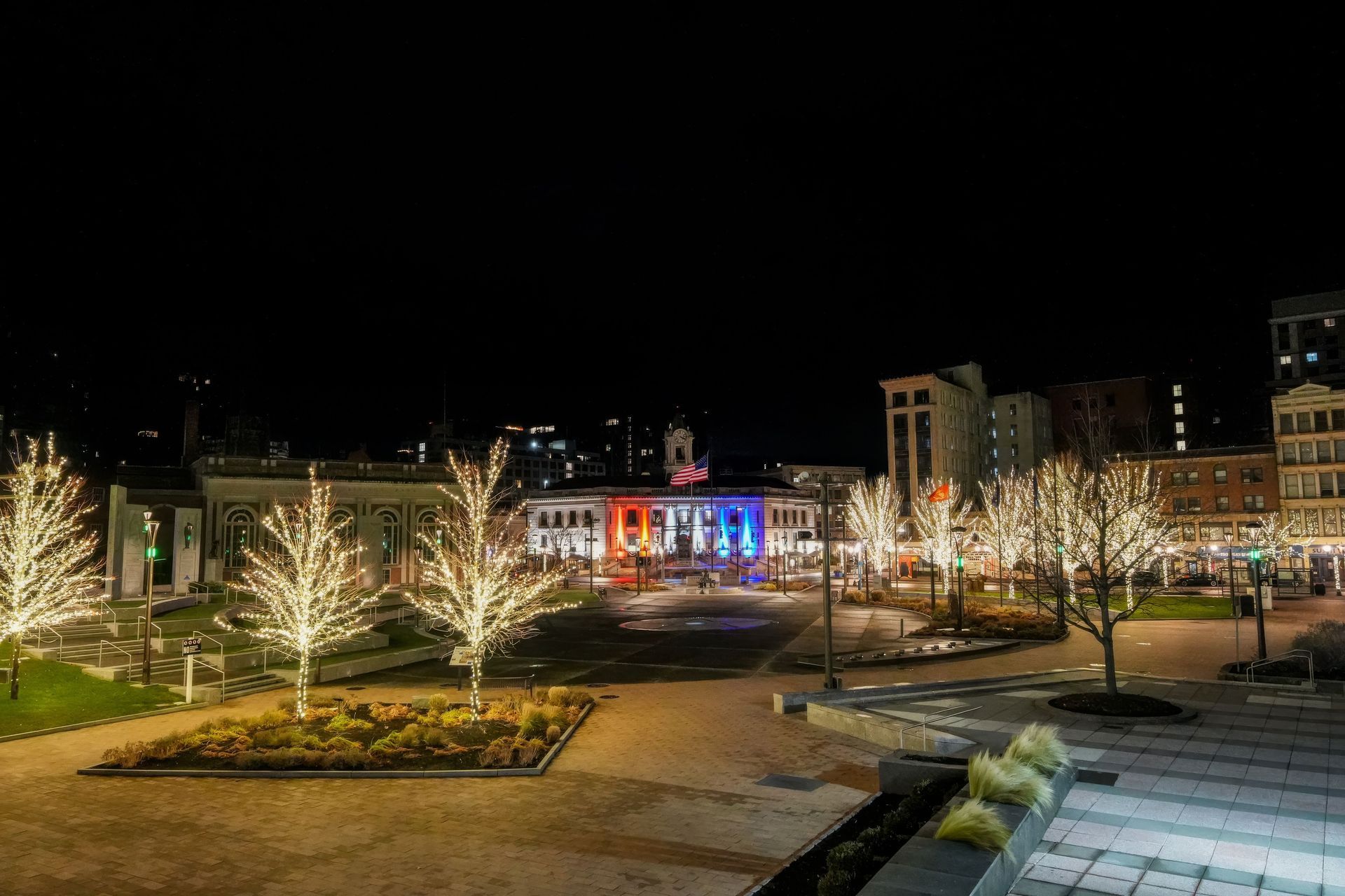 Illuminated town square at night with lit trees and colorful building lights