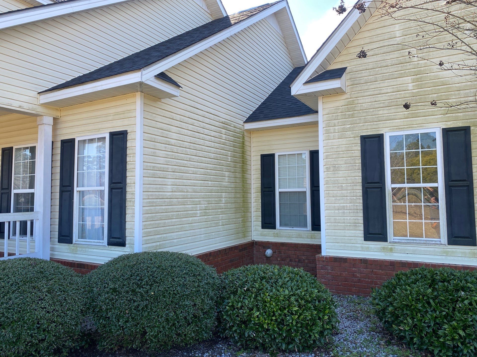 A yellow house with black shutters on the windows and bushes in front of it.