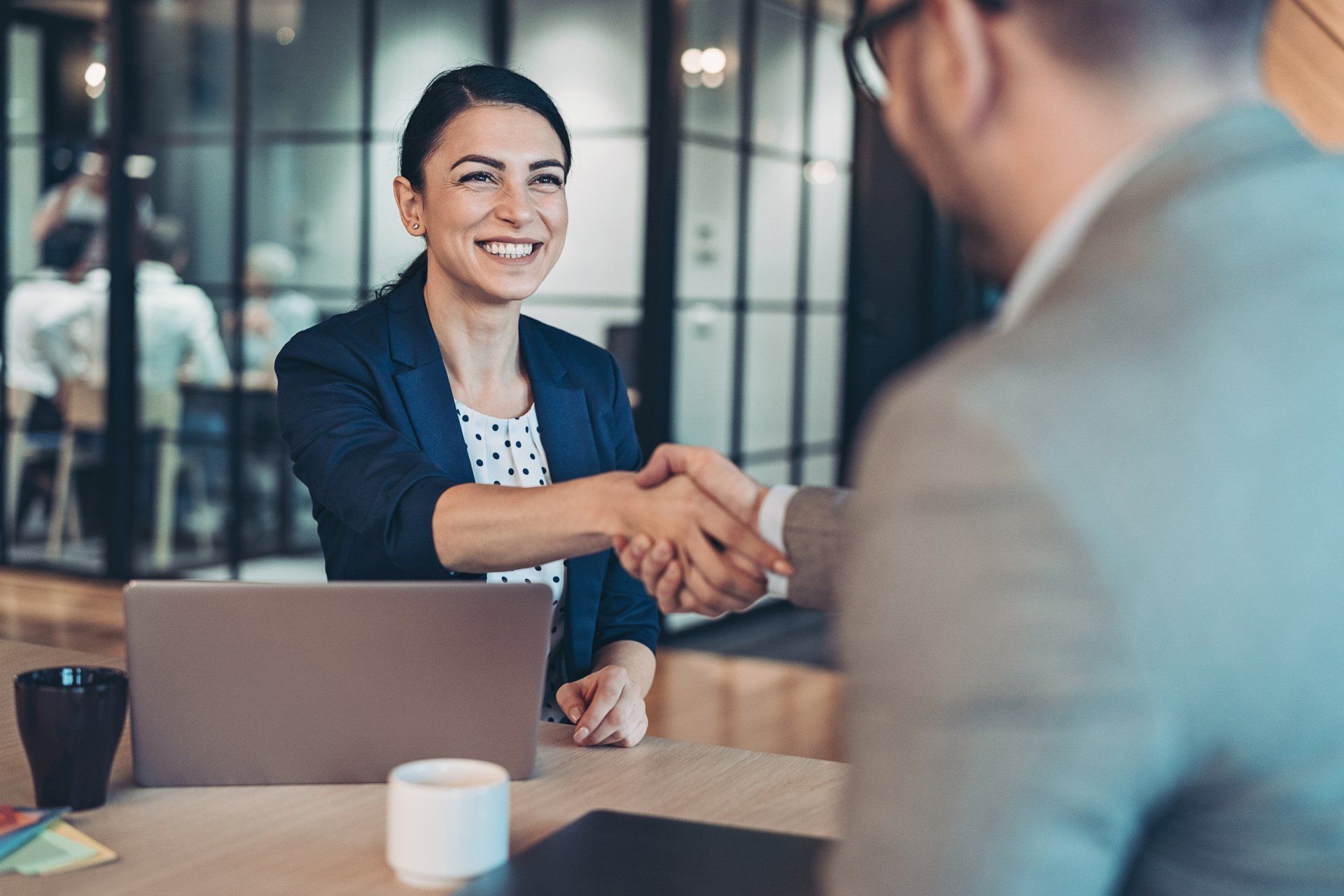 Woman in blazer shaking hands with a man, smiling in an office setting.