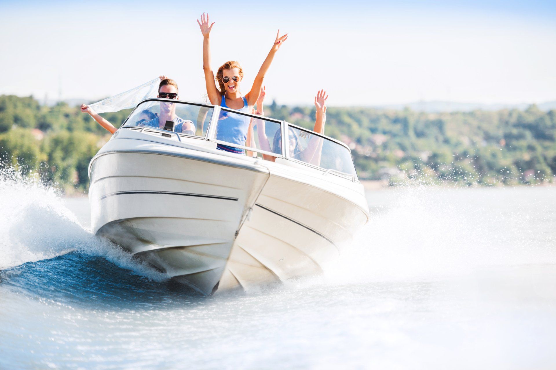 Three people on a speeding white motorboat, raising arms, on a sunny lake.