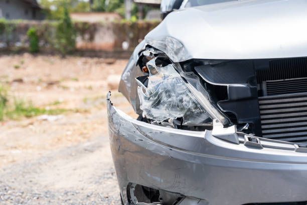 Damaged silver car front, headlight broken, dented fender, outdoor setting.