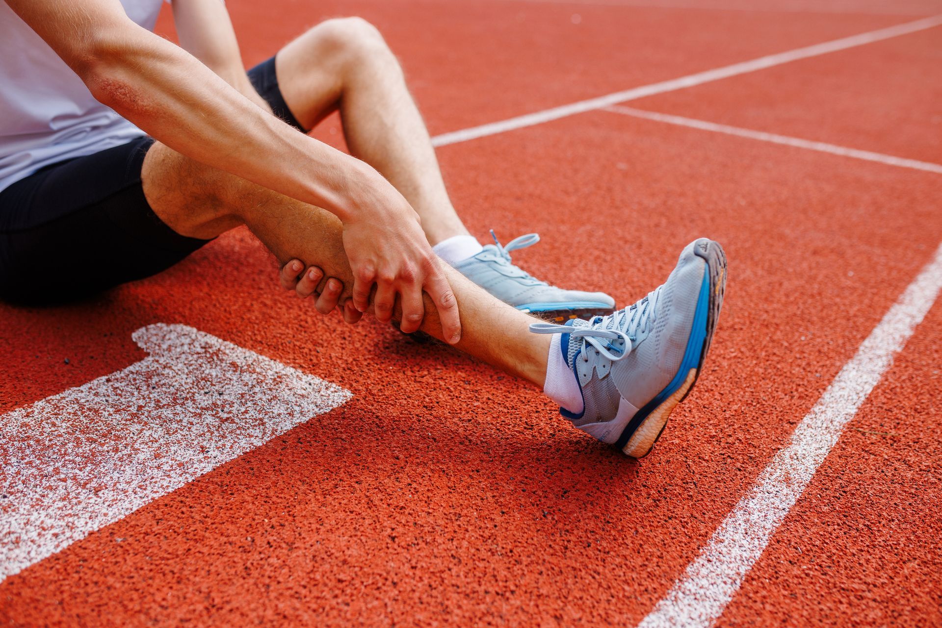 Runner on a red track clutching their ankle, possibly injured. Blue sneakers, white lines visible. Runner on a red track clutching their ankle, possibly injured. Blue sneakers, white lines visible.