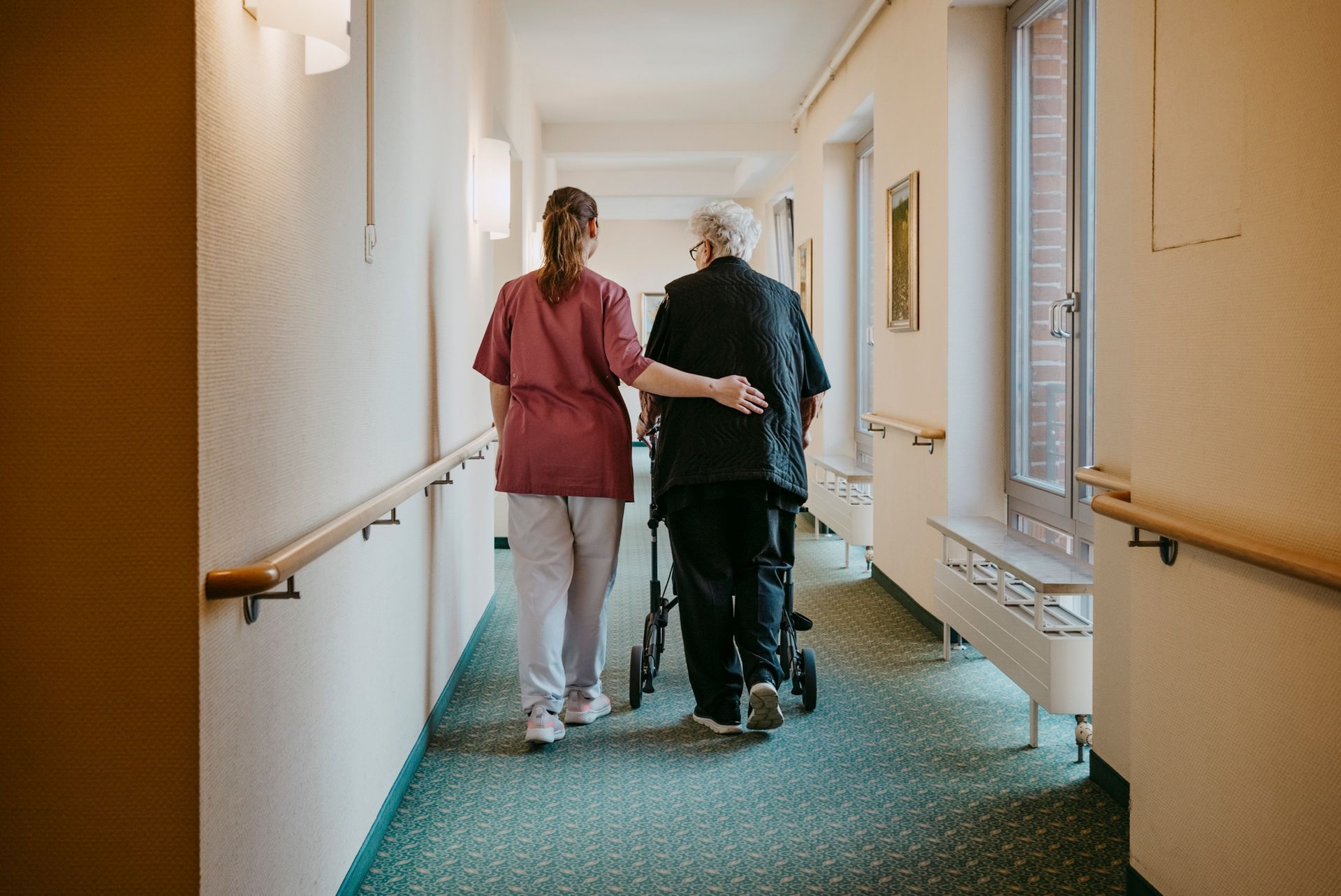A caregiver assists an elderly person using a walker down a long hallway with handrails.