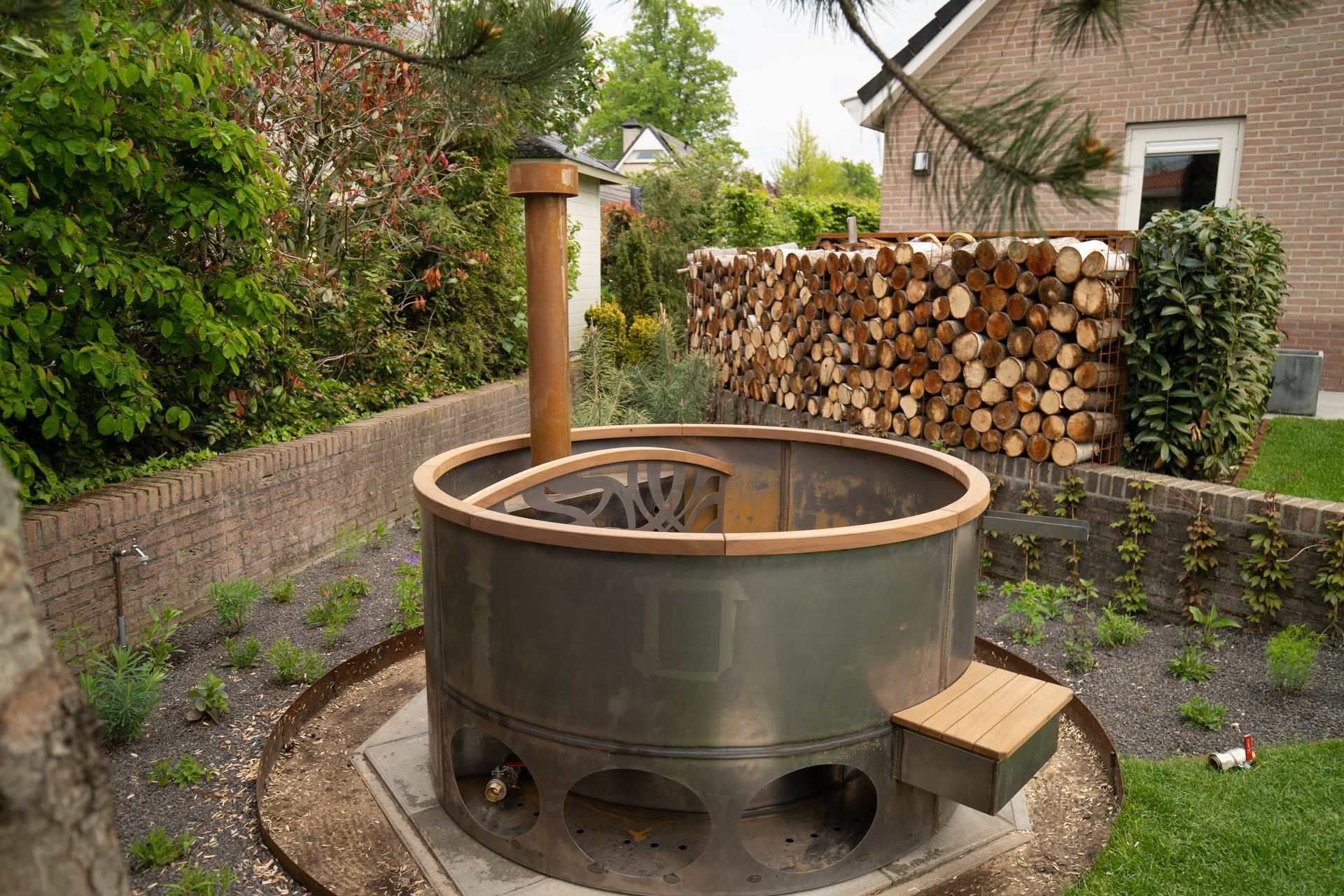 Bain à remous extérieur au feu de bois dans un cadre de jardin ; baignoire en acier gris, contour en bois, cheminée et bois de chauffage empilé à proximité.