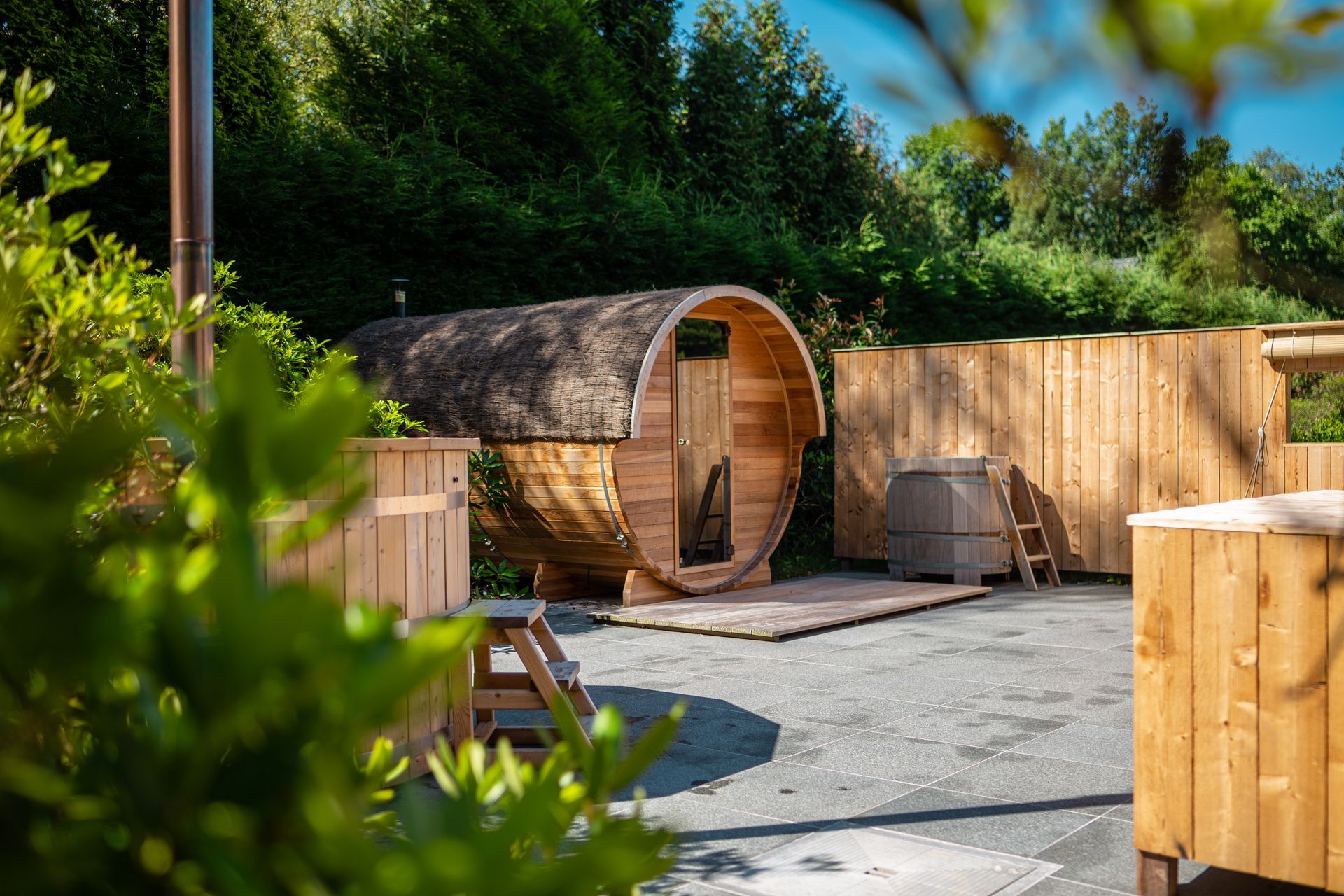Sauna en tonneau de bois à l'extérieur, entouré de structures en bois, d'arbres et d'arbustes.