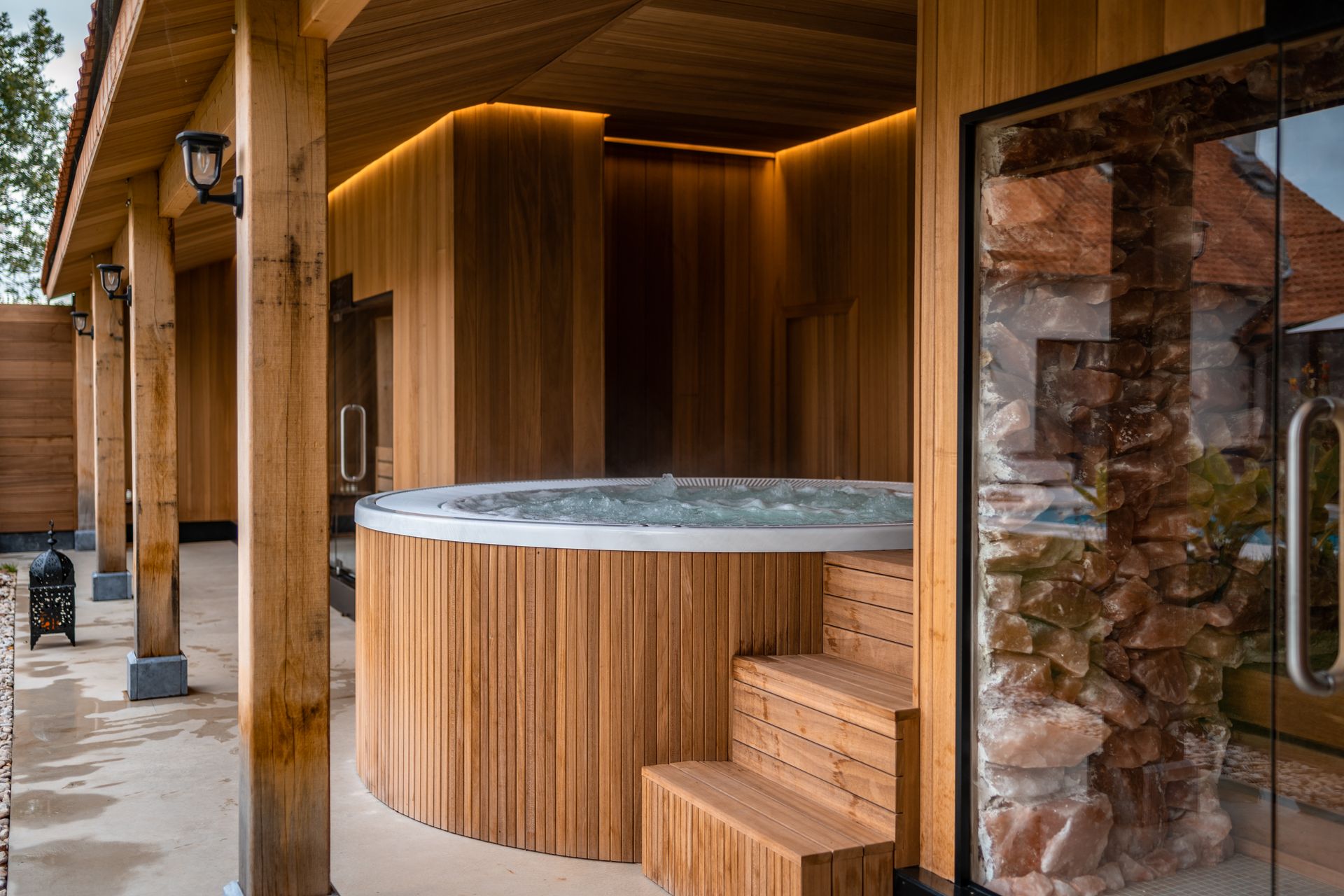 Spa en bois sur un patio, sous une structure en bois, avec marches menant au spa et porte vitrée.
