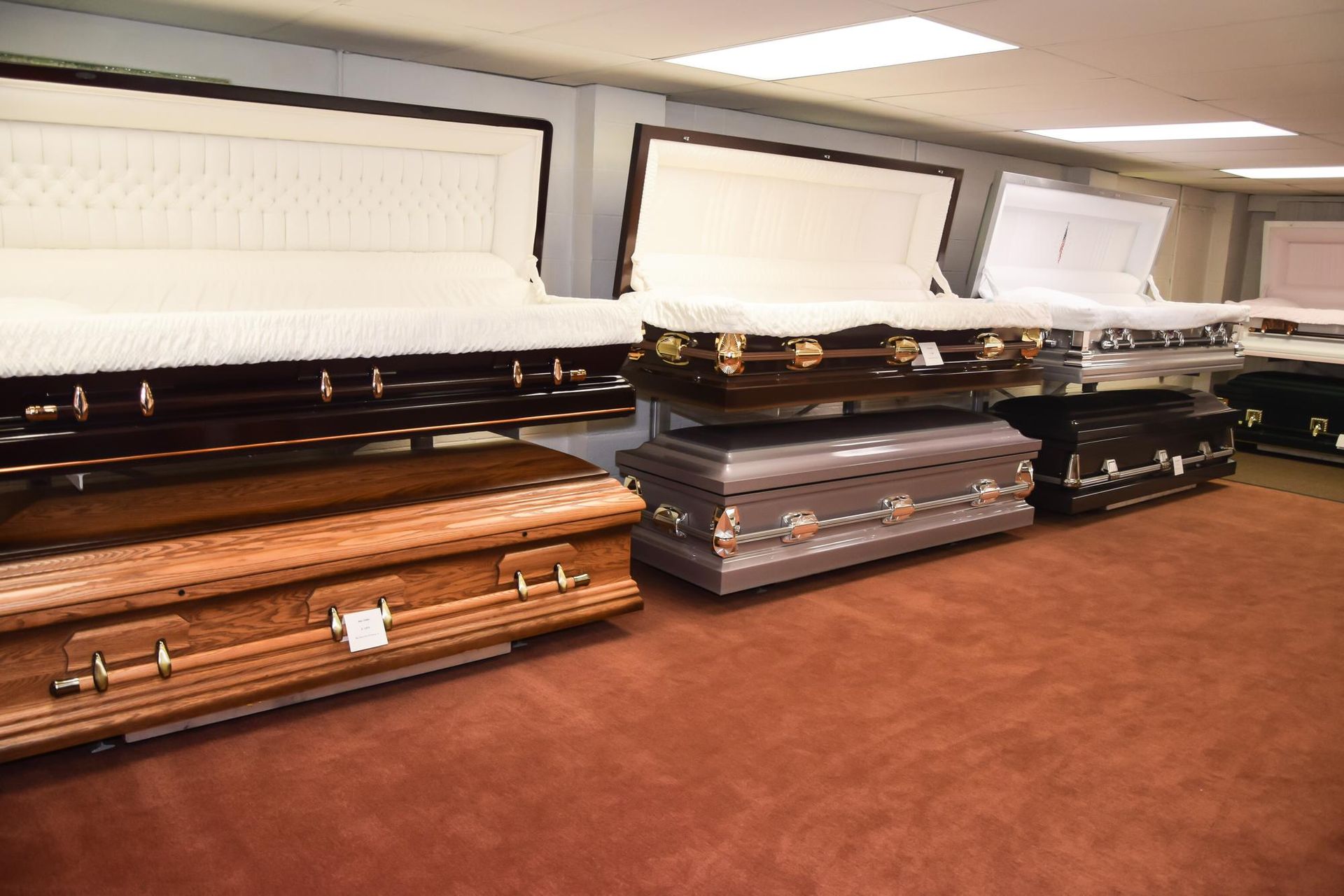 Several open coffins lined up in a funeral home room with white lids and brown carpet