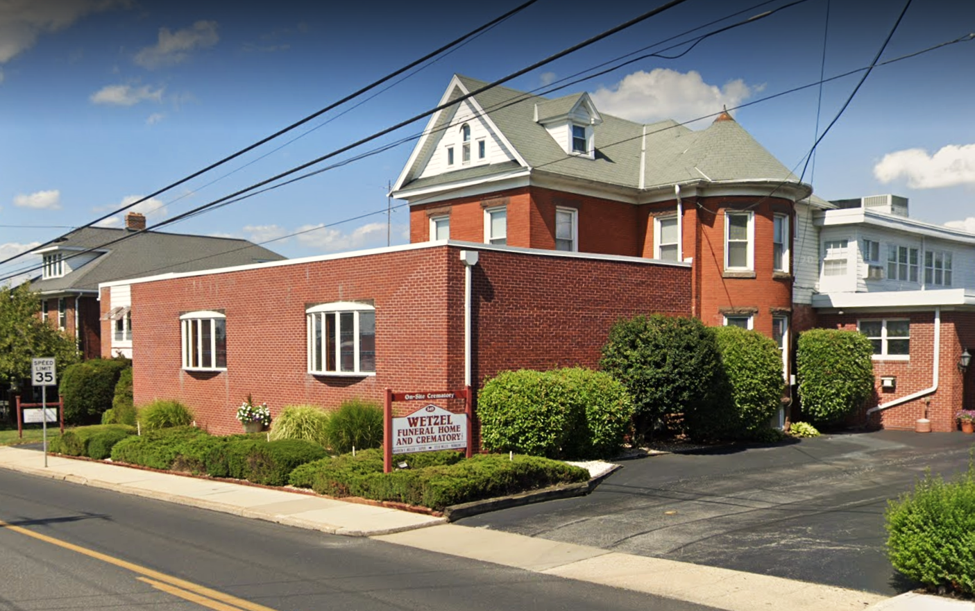 Red-brick church or civic building with a driveway, shrubs, and a street sign on a sunny corner.