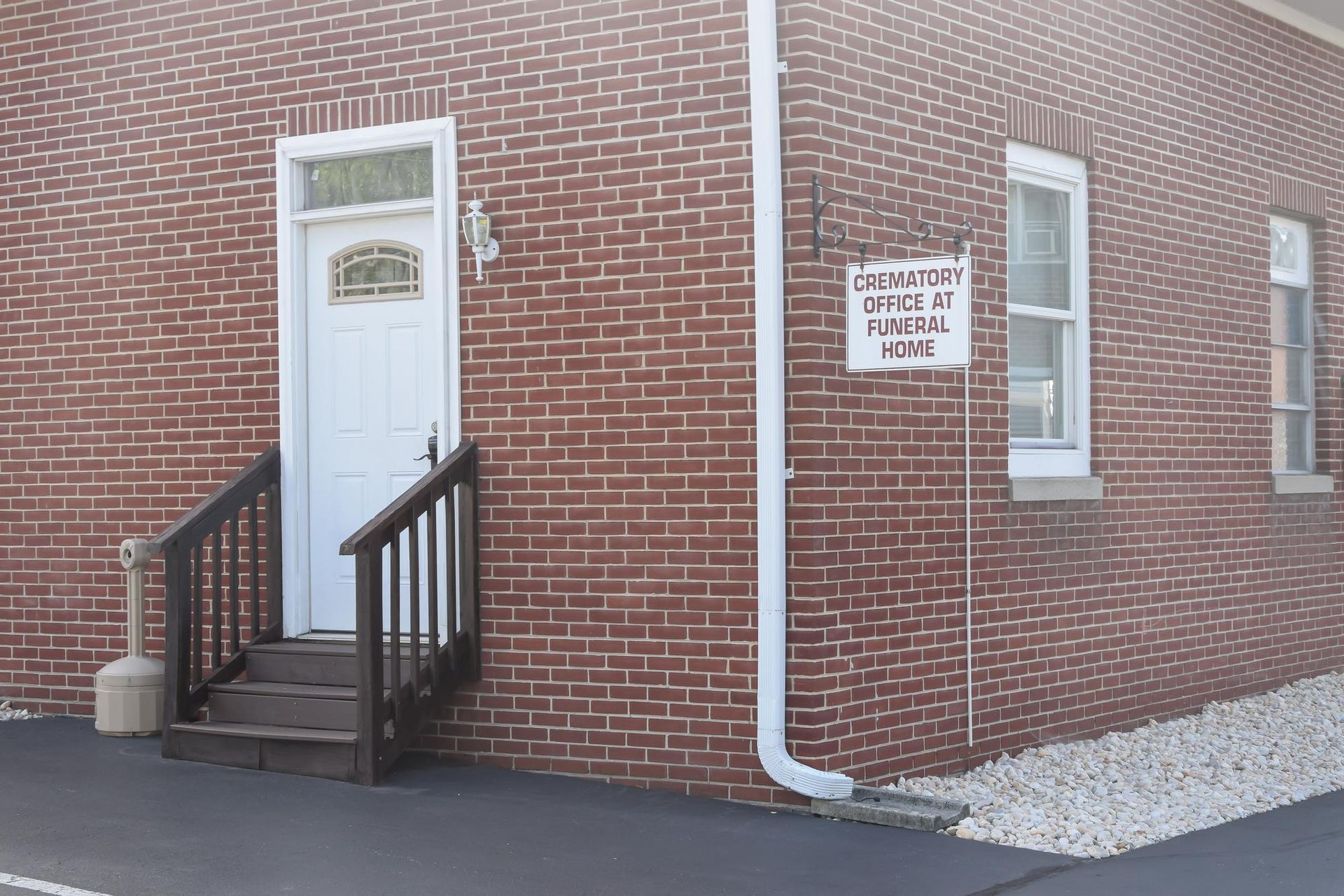 Brick building entrance with white door, black handrail, and a small warning sign by a window.