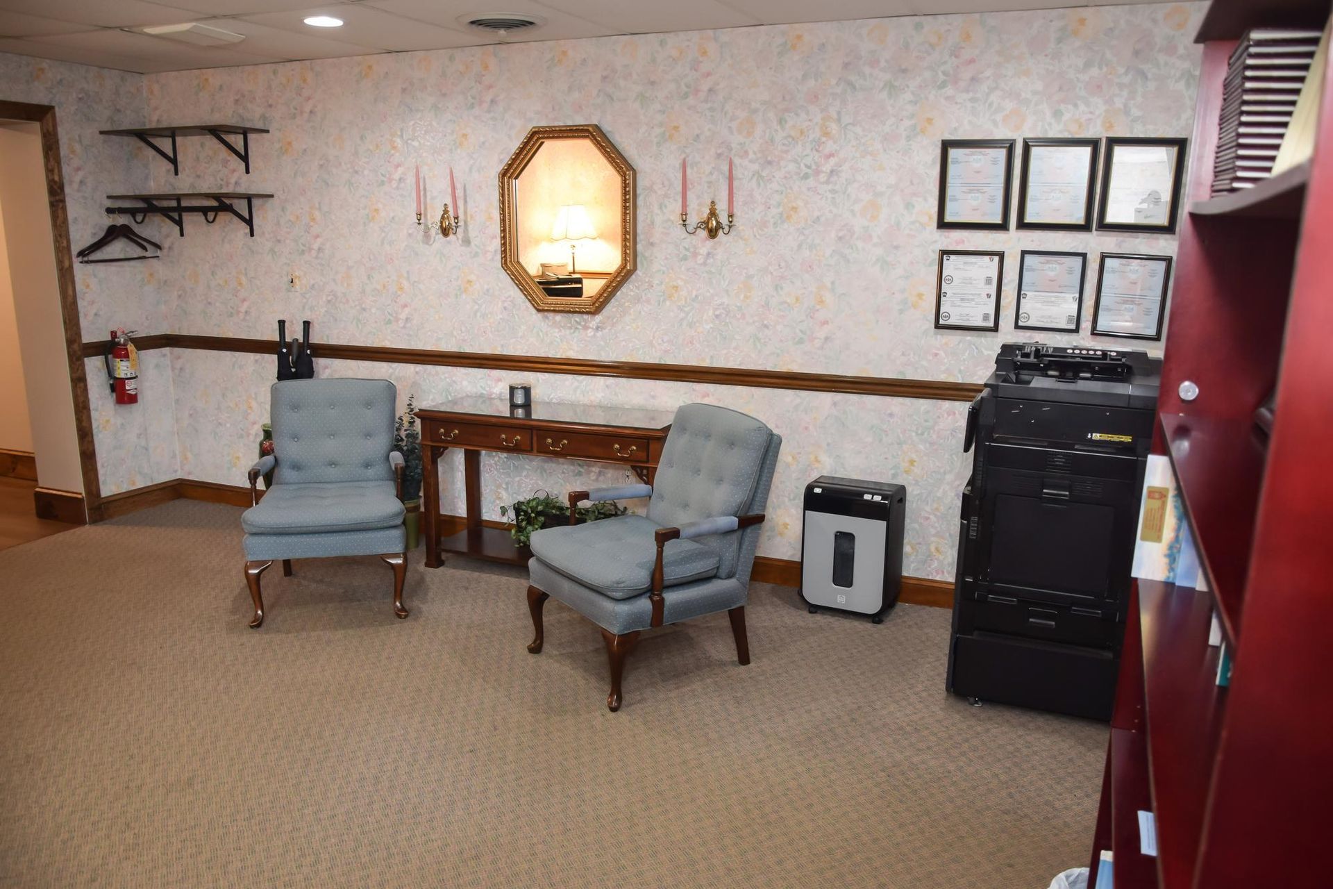 Waiting area with two blue chairs, small table, wall decor, and a copier beside shelves
