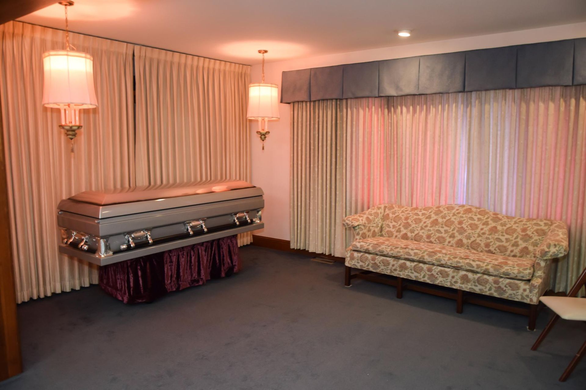 Funeral parlor room with a casket, soft pink lighting, and a patterned couch by curtained windows