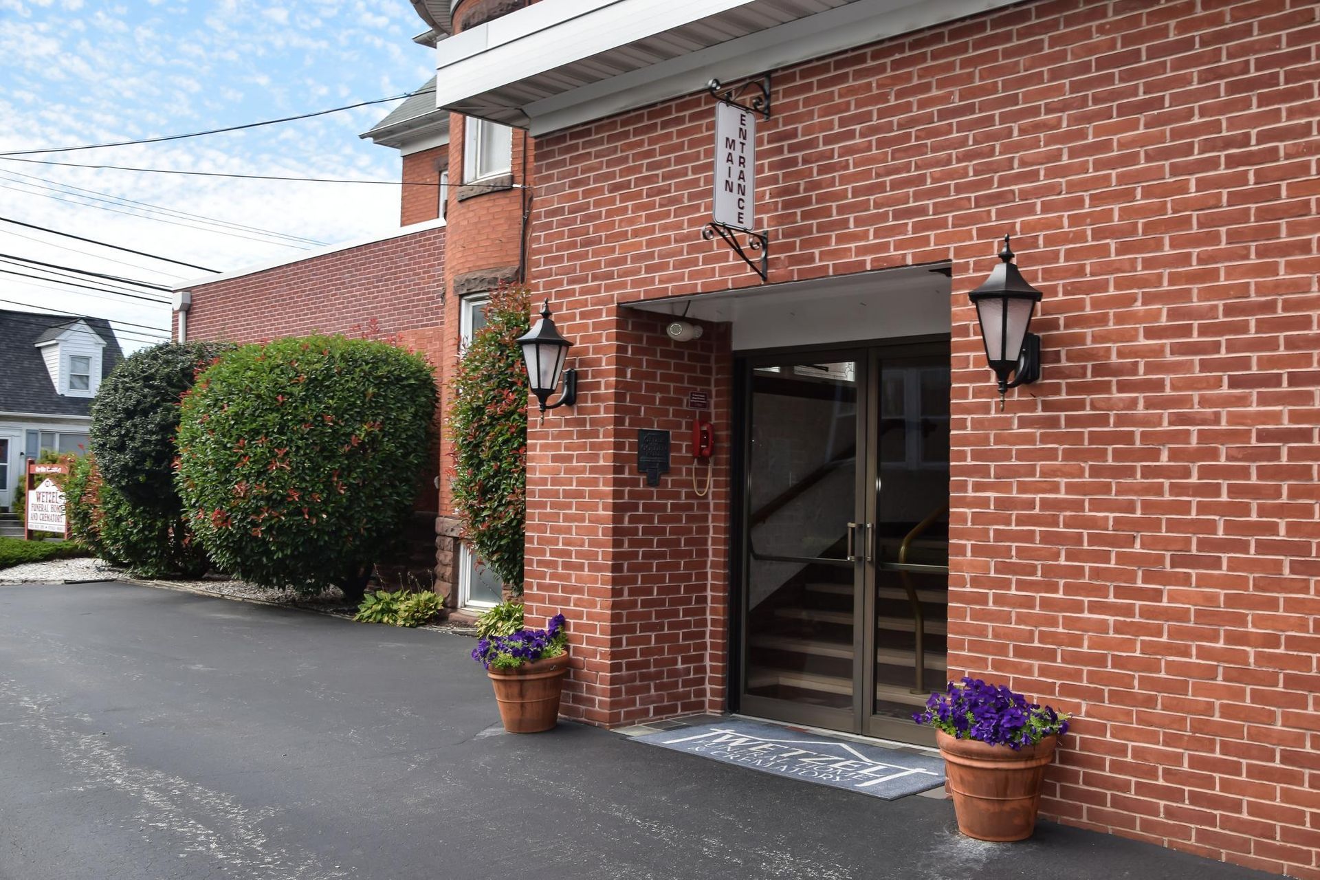Brick building entrance with glass door, wall lanterns, and flower pots by a paved driveway