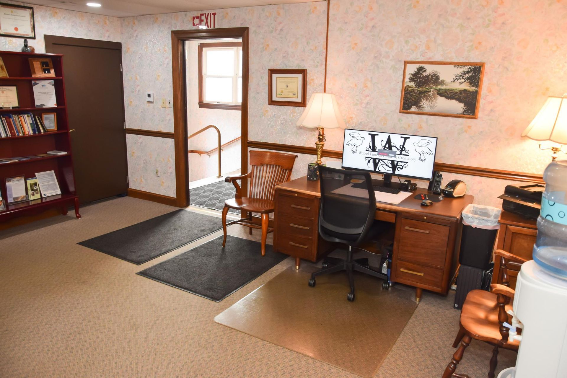 Office workspace with wooden desks, computer monitors, lamps, and chairs on carpeted floor