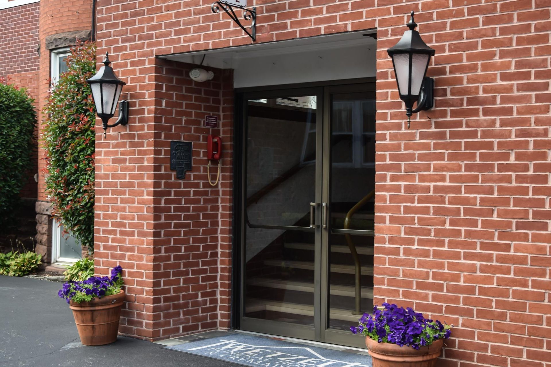 Brick building entrance with glass door, black lanterns, and flower pots by the doorway