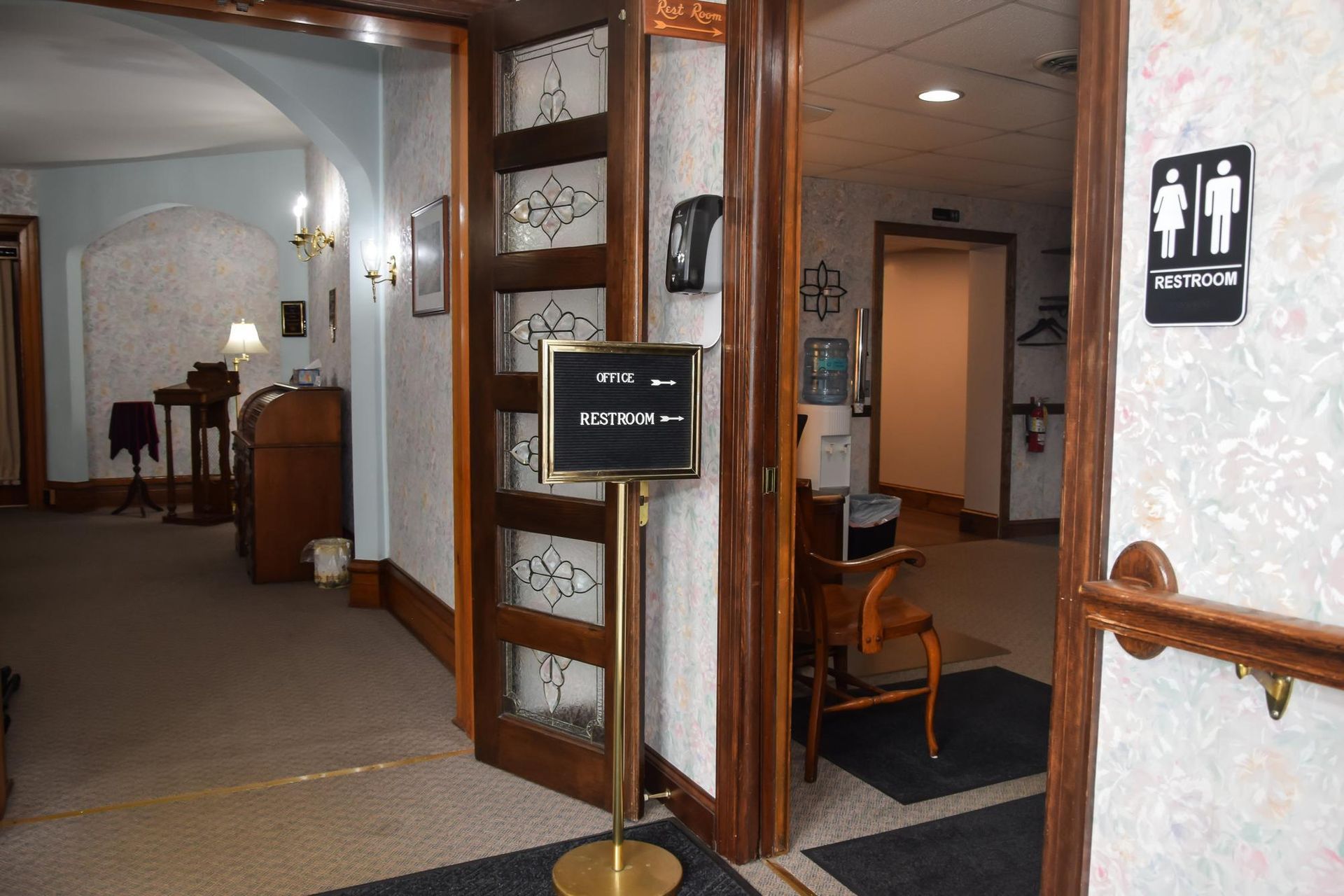 Interior hallway entrance with wooden doors, a sign stand, and chairs in a carpeted building lobby