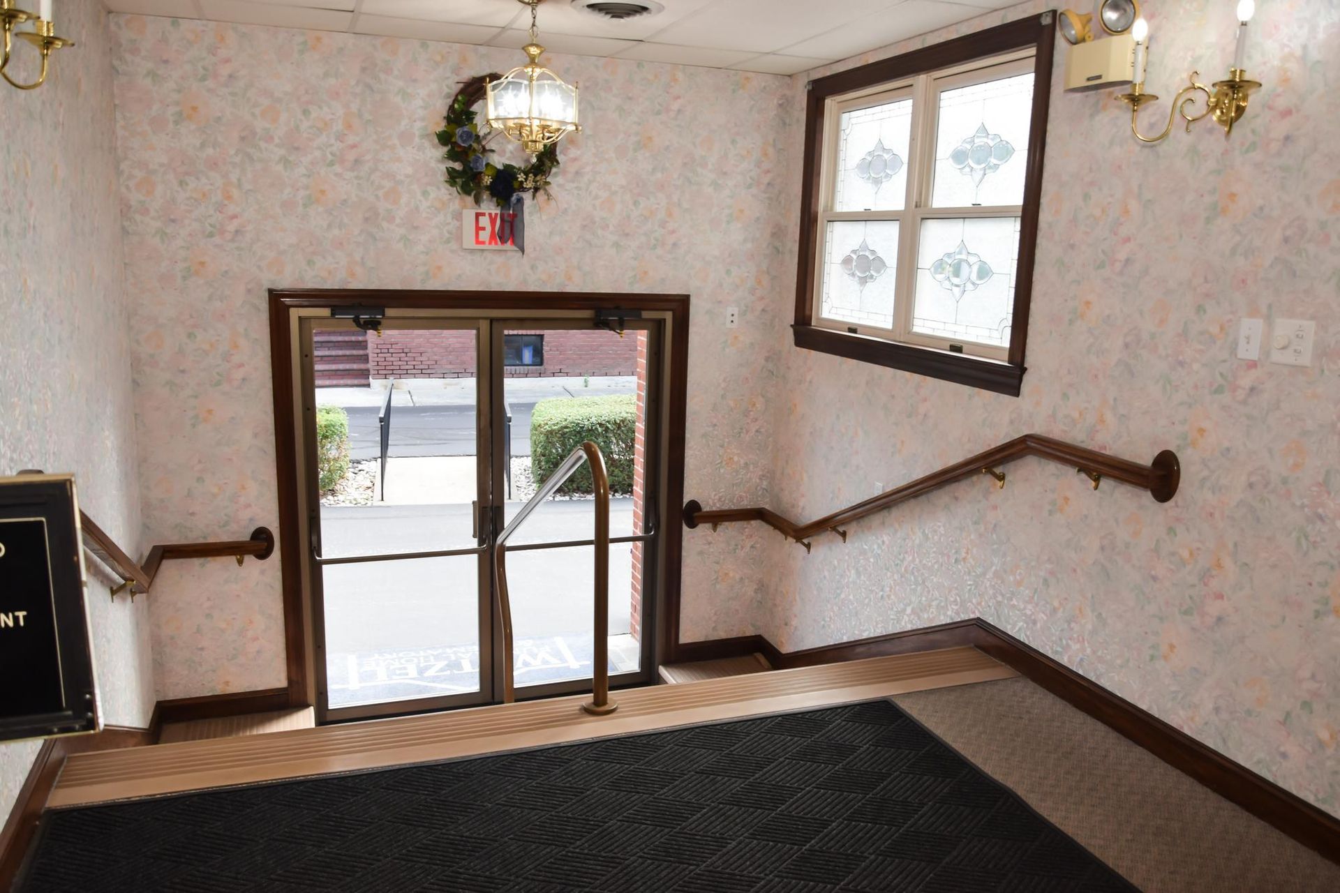 Stair landing with patterned wallpaper, hanging lights, wooden handrails, and glass doors to the street.
