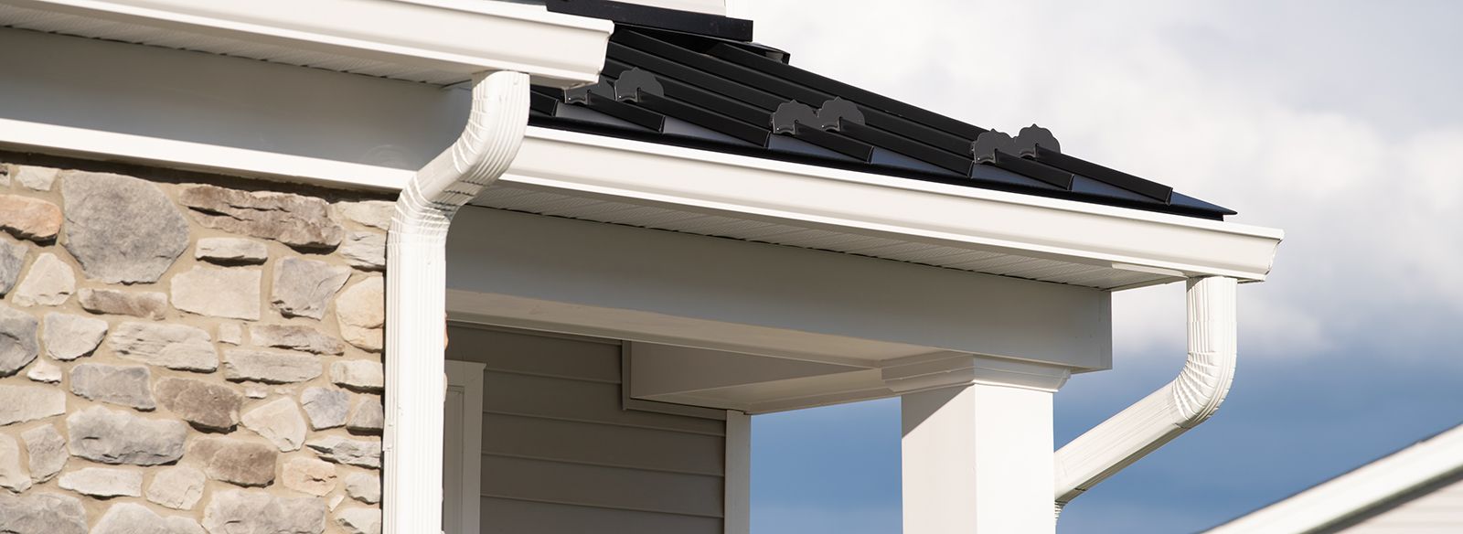 White gutters on a house with stone siding and black roof tiles against a cloudy sky.