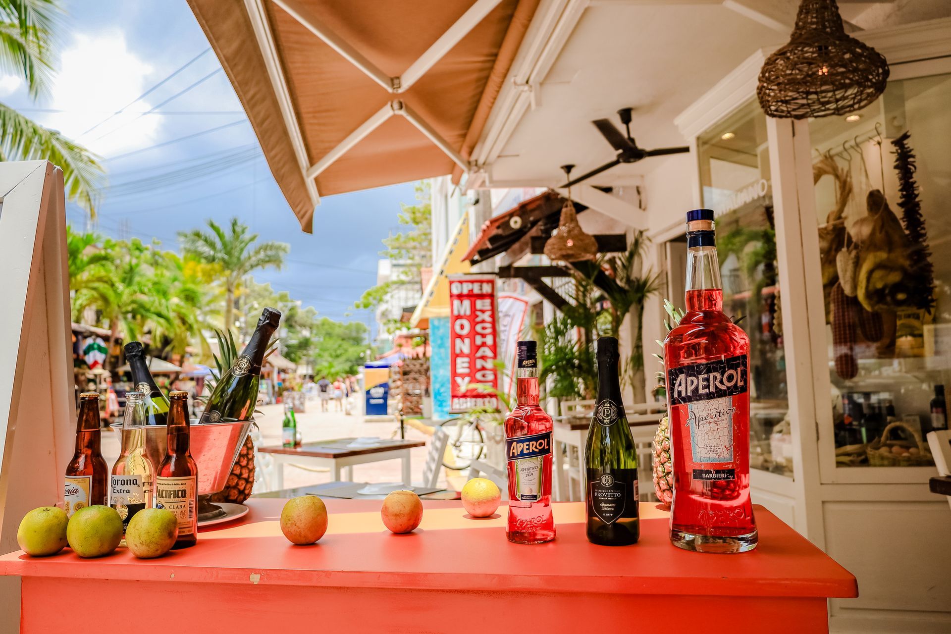 Red bar with liquor bottles, apples, palm trees, and people walking in a tropical outdoor setting.