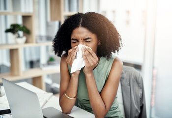 A woman is blowing her nose while sitting at a desk with a laptop.