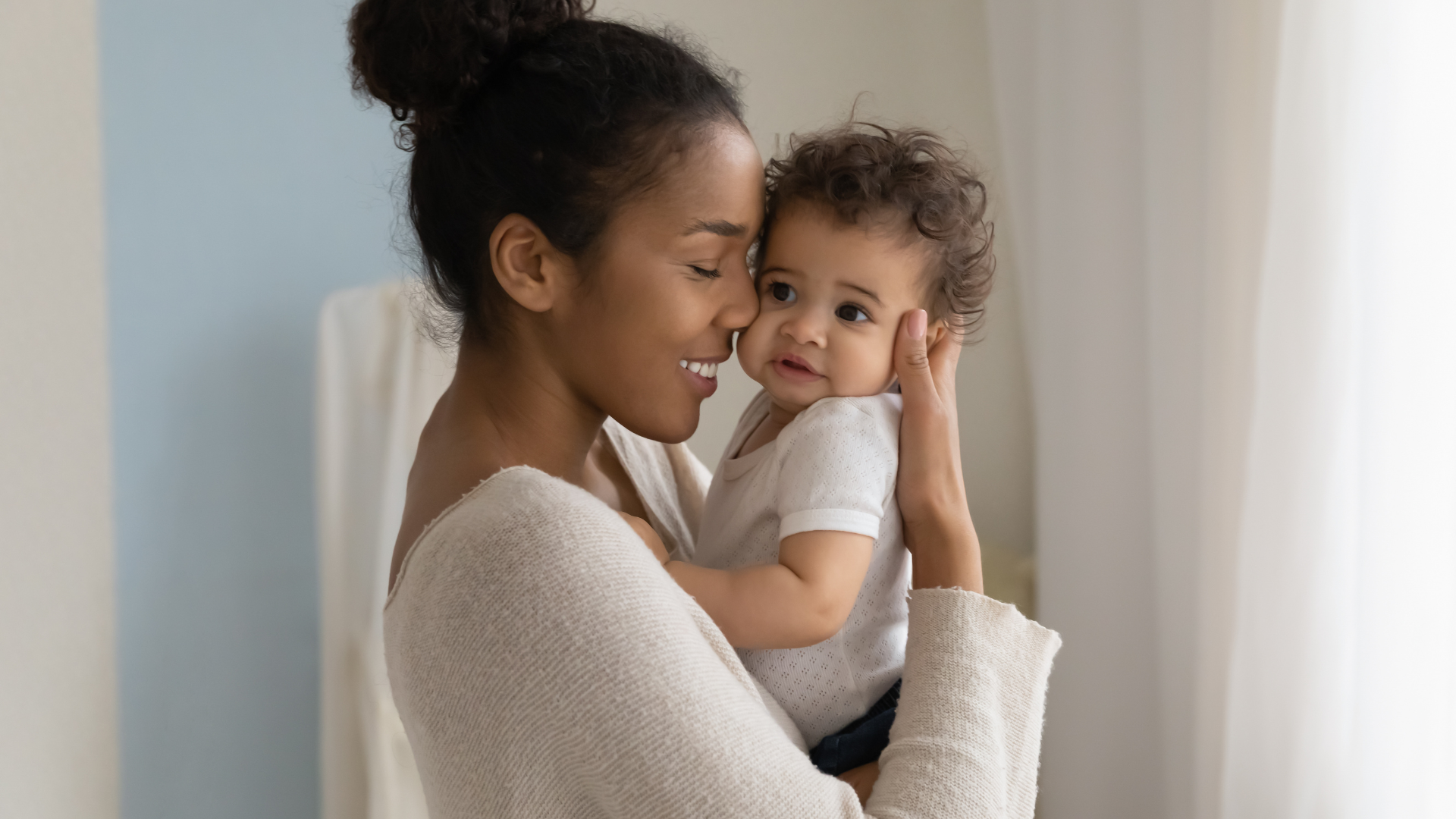 Woman holding baby, smiling, near window. Baby looks at the woman lovingly.