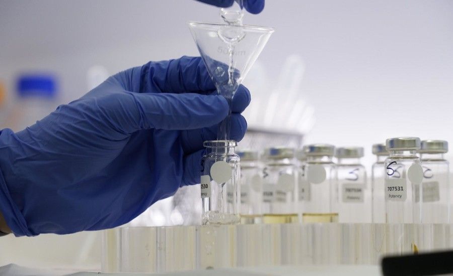 A gloved hand pours liquid through a funnel into a small vial in a lab setting.