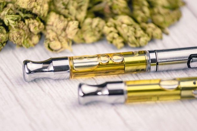Two vape cartridges filled with yellow liquid, with cannabis buds in the background.