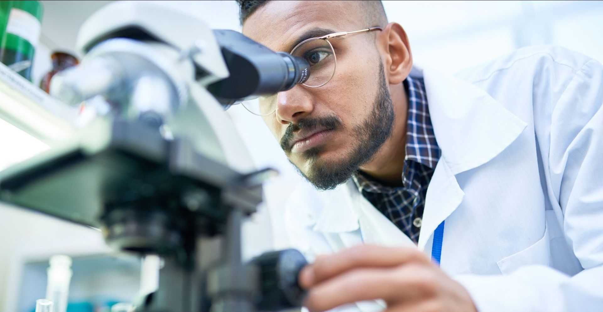 A person in a lab coat looks through a microscope, adjusting a knob.