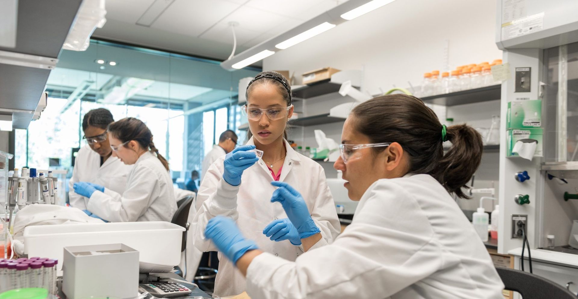 Scientists in lab coats, gloves, and safety glasses, working with equipment in a laboratory.