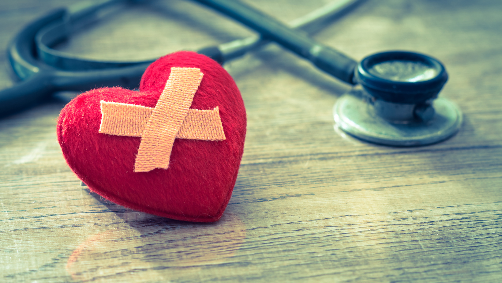 A red heart with a cross on it is next to a stethoscope on a wooden table.