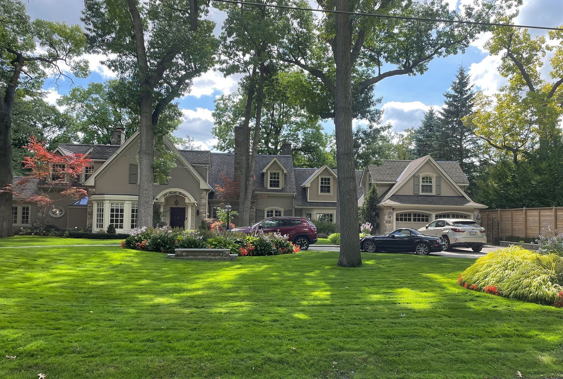 Large house with multiple gabled sections, surrounded by a green lawn and trees, vehicles parked in the driveway.