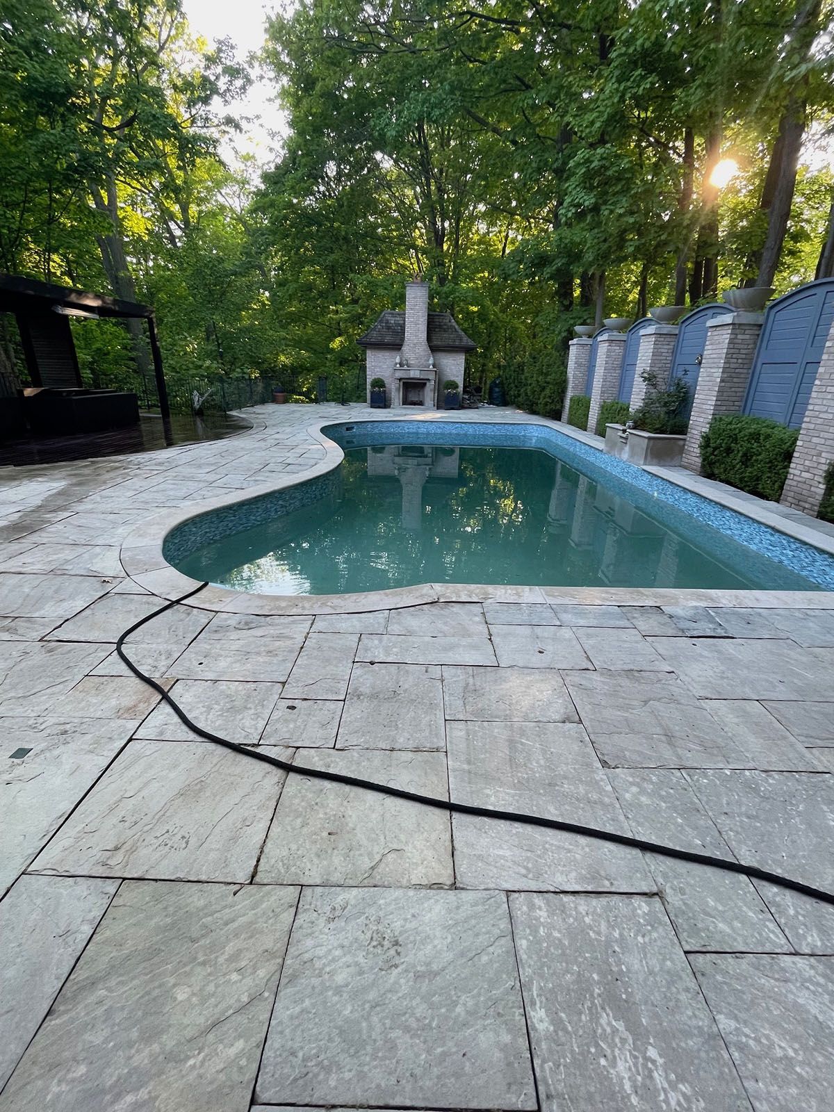 Pool with stone deck, small fireplace, trees in background, and sunlight.