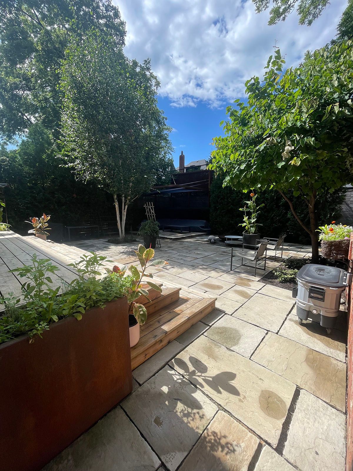 Patio with stone pavers, steps, trees, and plants. Cloudy sky overhead.