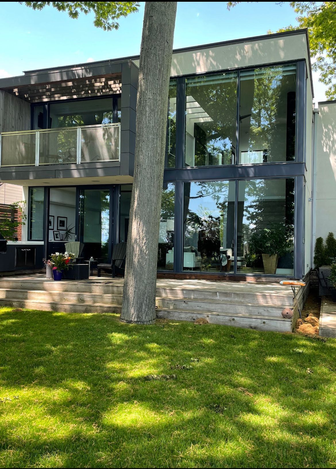 Modern two-story house with large glass windows overlooking a lake. A tree is in the foreground.