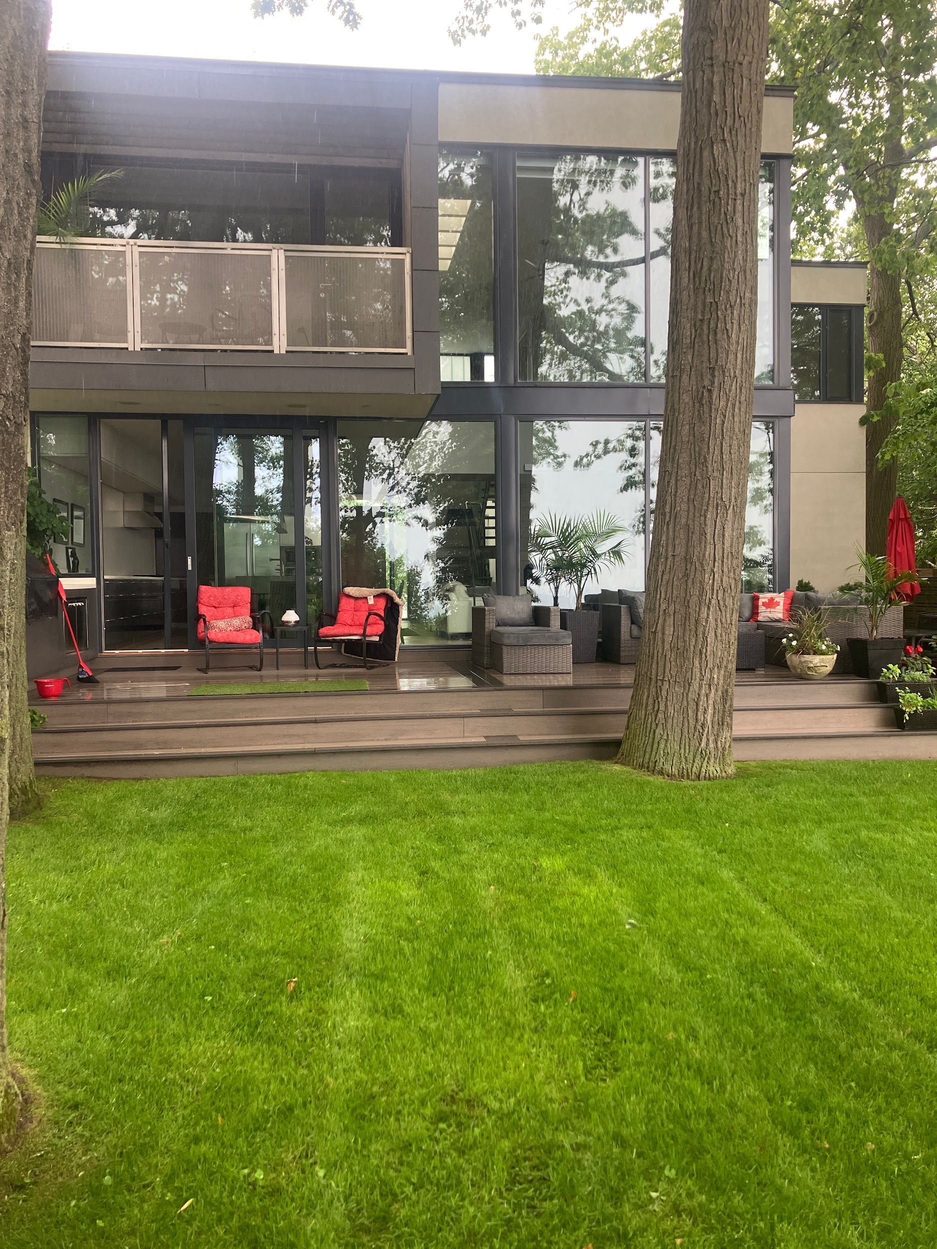 Modern house with large windows and deck, red chairs on patio, green lawn.