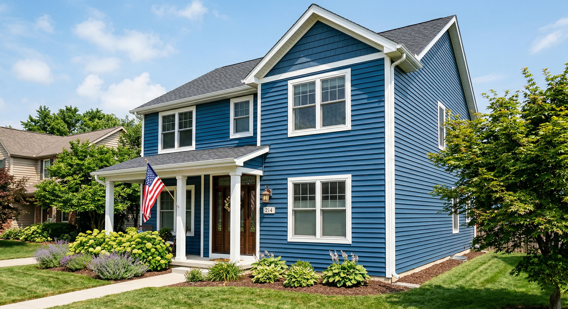Blue two-story house with white trim, front porch, and an American flag amid a landscaped yard.
