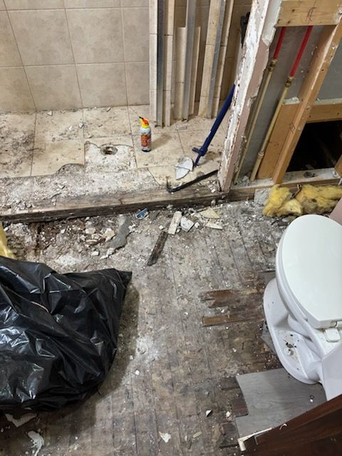 Damaged bathroom floor with exposed debris, toilet on right, and black trash bag on the left
